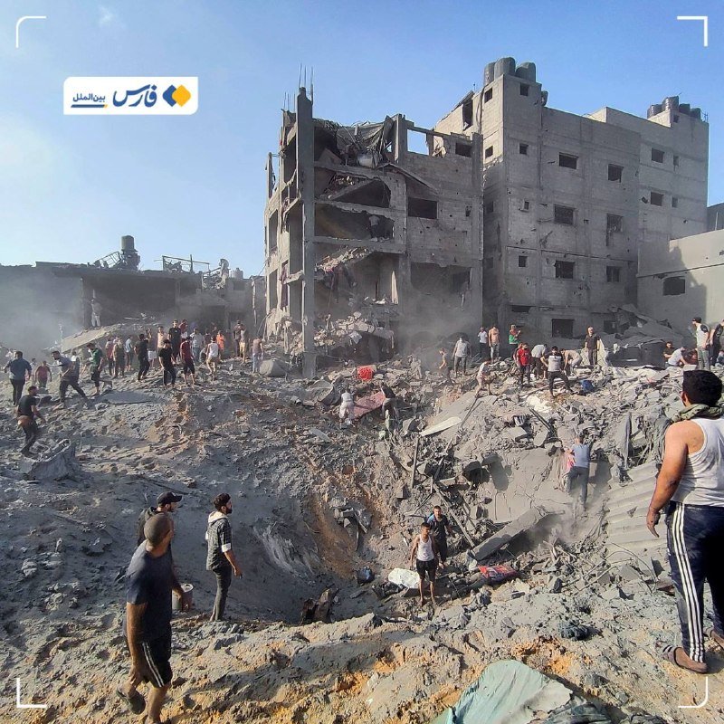 People gather amid the rubble of heavily damaged and partially collapsed concrete buildings under a clear sky, with a large crater visible in the foreground debris.