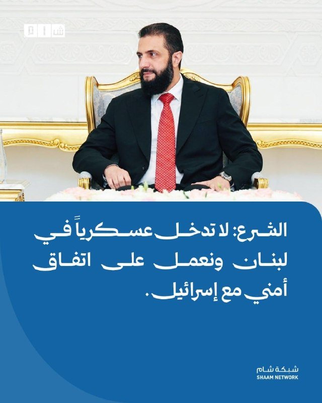 A bearded man in a black suit and red tie sits in an ornate chair against a white wall, with Arabic text and the "Shaam Network" logo displayed below.