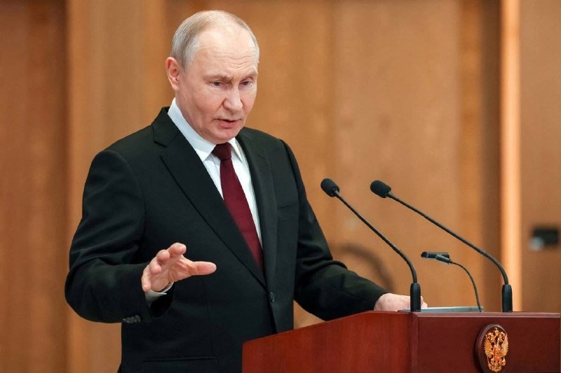 A gray-haired man in a black suit and red tie speaks at a wooden podium bearing a gold double-headed eagle emblem, gesturing with one hand.
