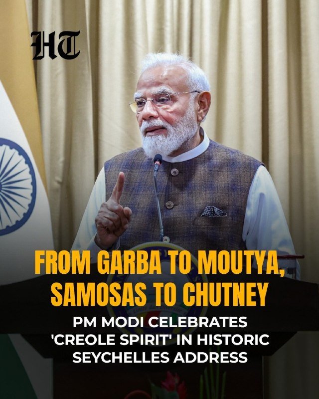 A man with a white beard and glasses speaks at a podium with a microphone, gesturing with his index finger raised, beside an Indian flag.