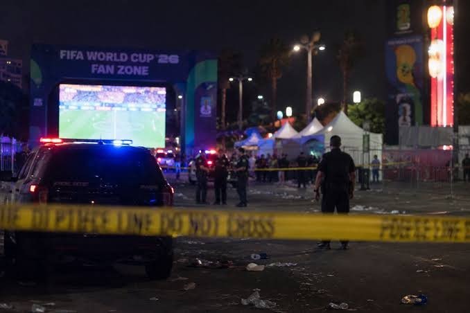 Police officers stand behind yellow police tape near a dark SUV outside a "FIFA World Cup '26 Fan Zone" venue at night, with debris scattered on the ground.