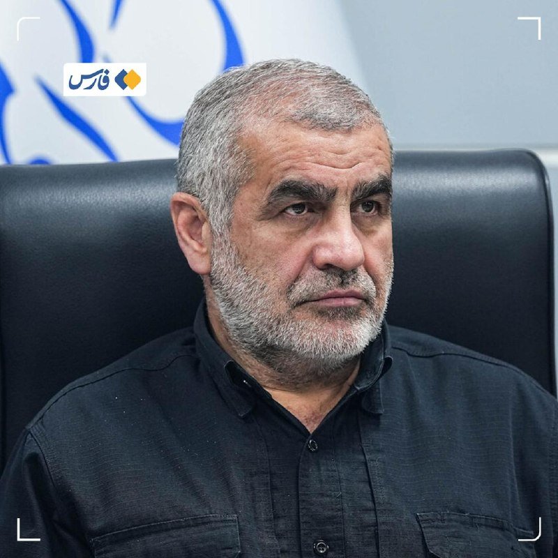 A gray-bearded man in a black shirt sits in a black office chair, photographed in close-up against a blurred background featuring a blue and white graphic.
