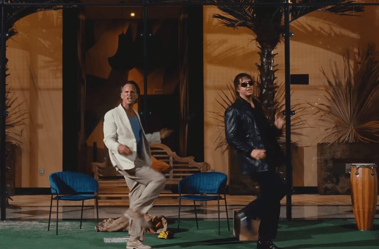 Two men dance energetically on a stage with blue chairs, palm plants, and a conga drum visible in the background.