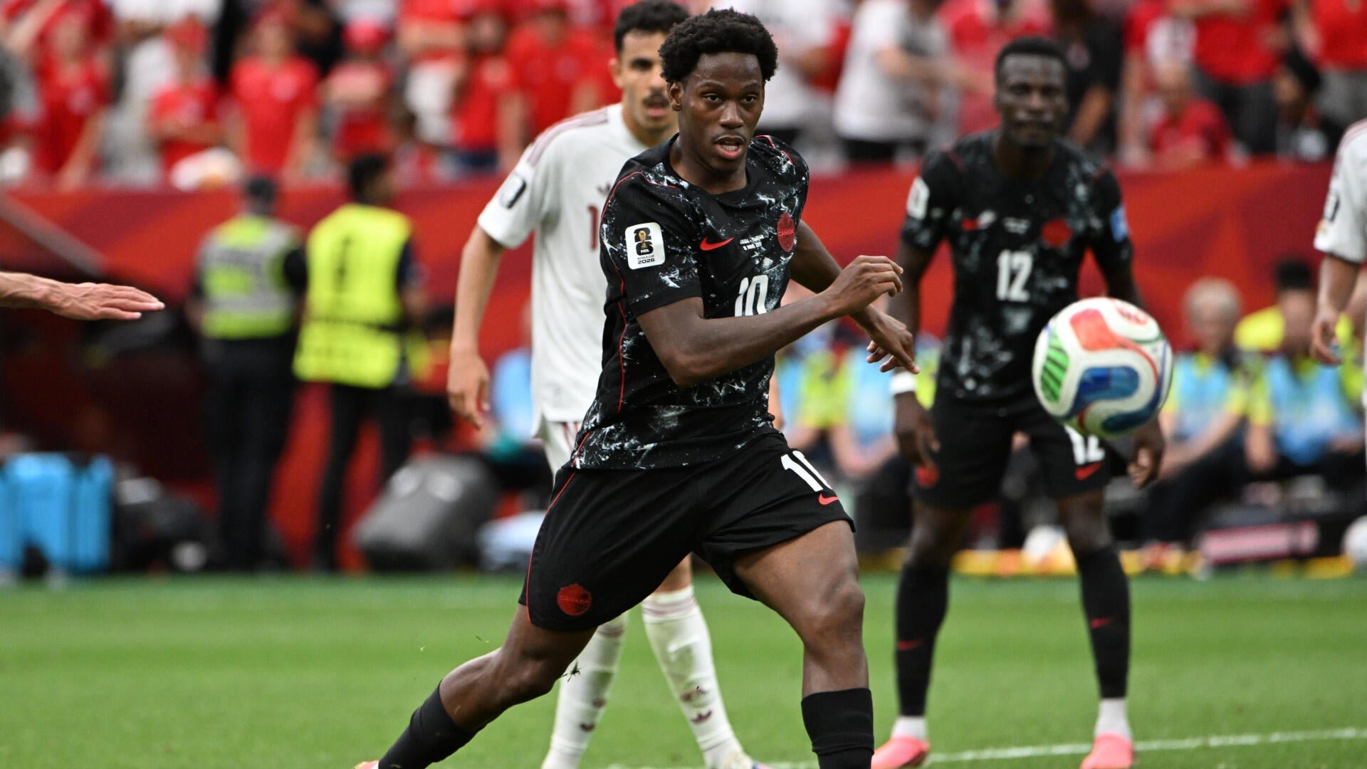 A soccer player in a black jersey (#10) runs toward a ball mid-air during a match, with another teammate (#12) and an opposing player in white visible behind him on the pitch.