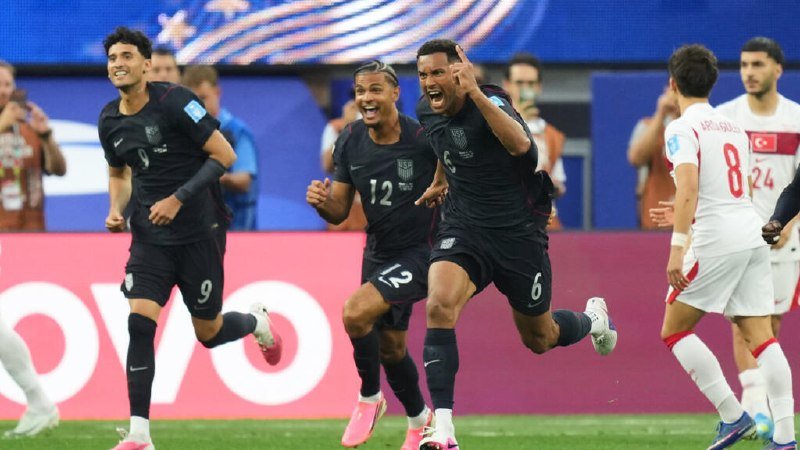 Three soccer players in dark jerseys celebrate exuberantly on the field while opposing players in white and red jerseys walk away dejectedly.