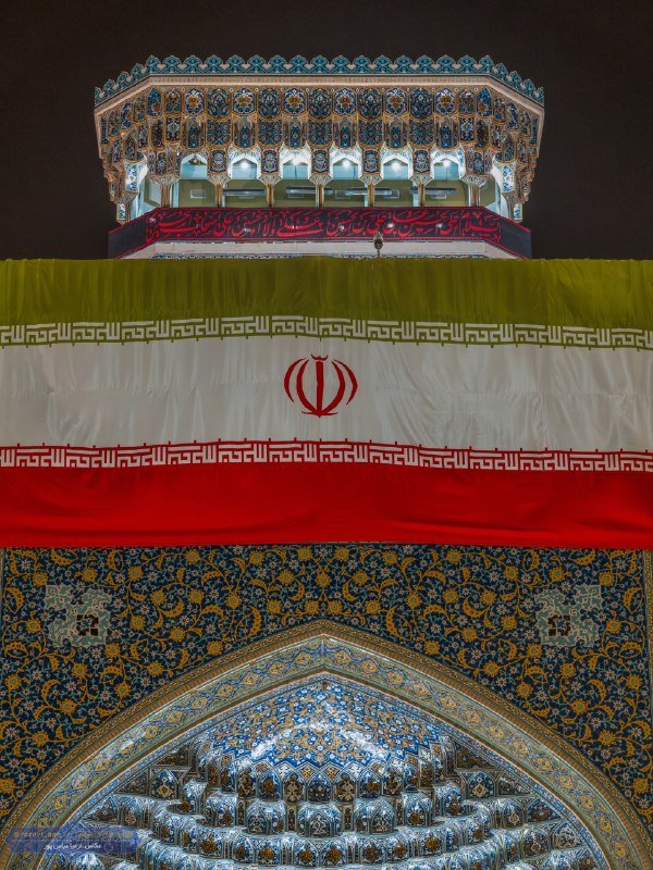 An illuminated ornate shrine with blue tilework is draped with a large Iranian flag bearing the national emblem at night.