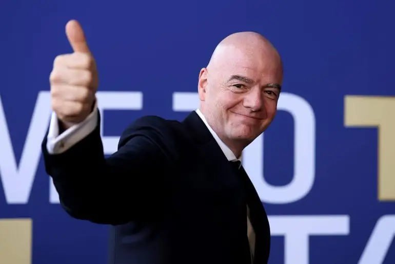 A bald man in a dark suit and tie smiles while giving a thumbs-up gesture against a blue backdrop with partial white and yellow lettering.