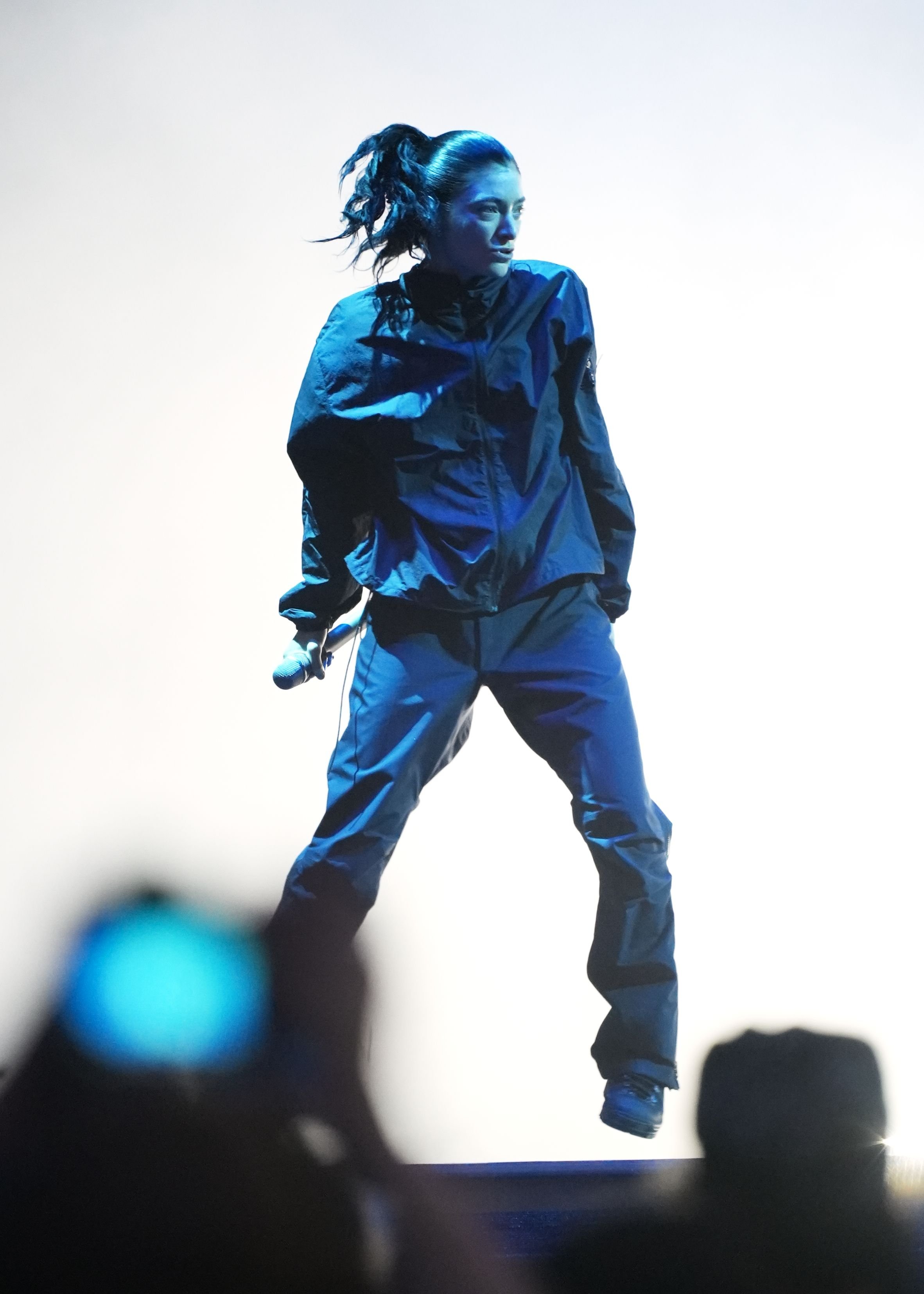 A performer in a dark jacket and pants jumps mid-air while holding a microphone, illuminated by bright blue stage lighting.