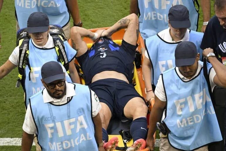 Medical staff wearing FIFA Medical vests carry an injured soccer player on a stretcher across a grass field.