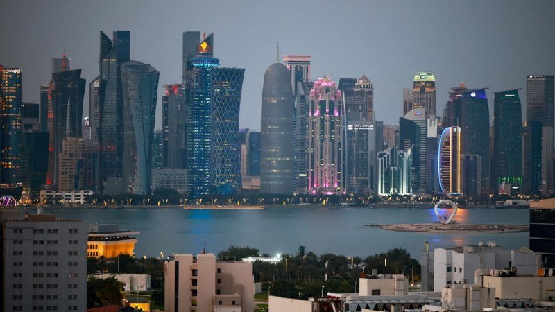 A skyline of illuminated skyscrapers rises across a body of water at dusk, with low-rise buildings and greenery visible in the foreground.