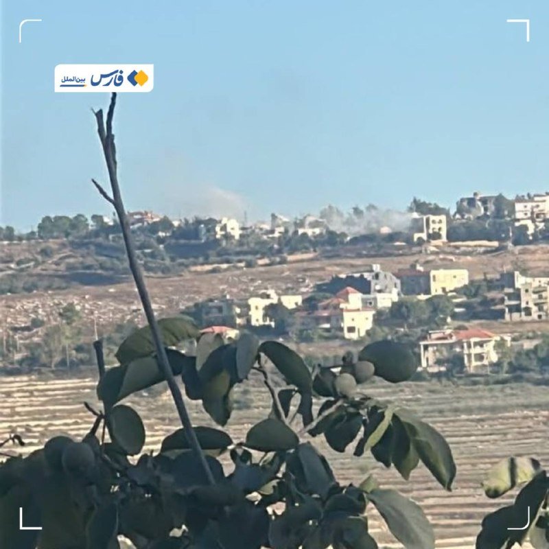A distant view across a terraced hillside shows light-colored buildings with plumes of smoke rising, framed by leafy branches in the foreground.