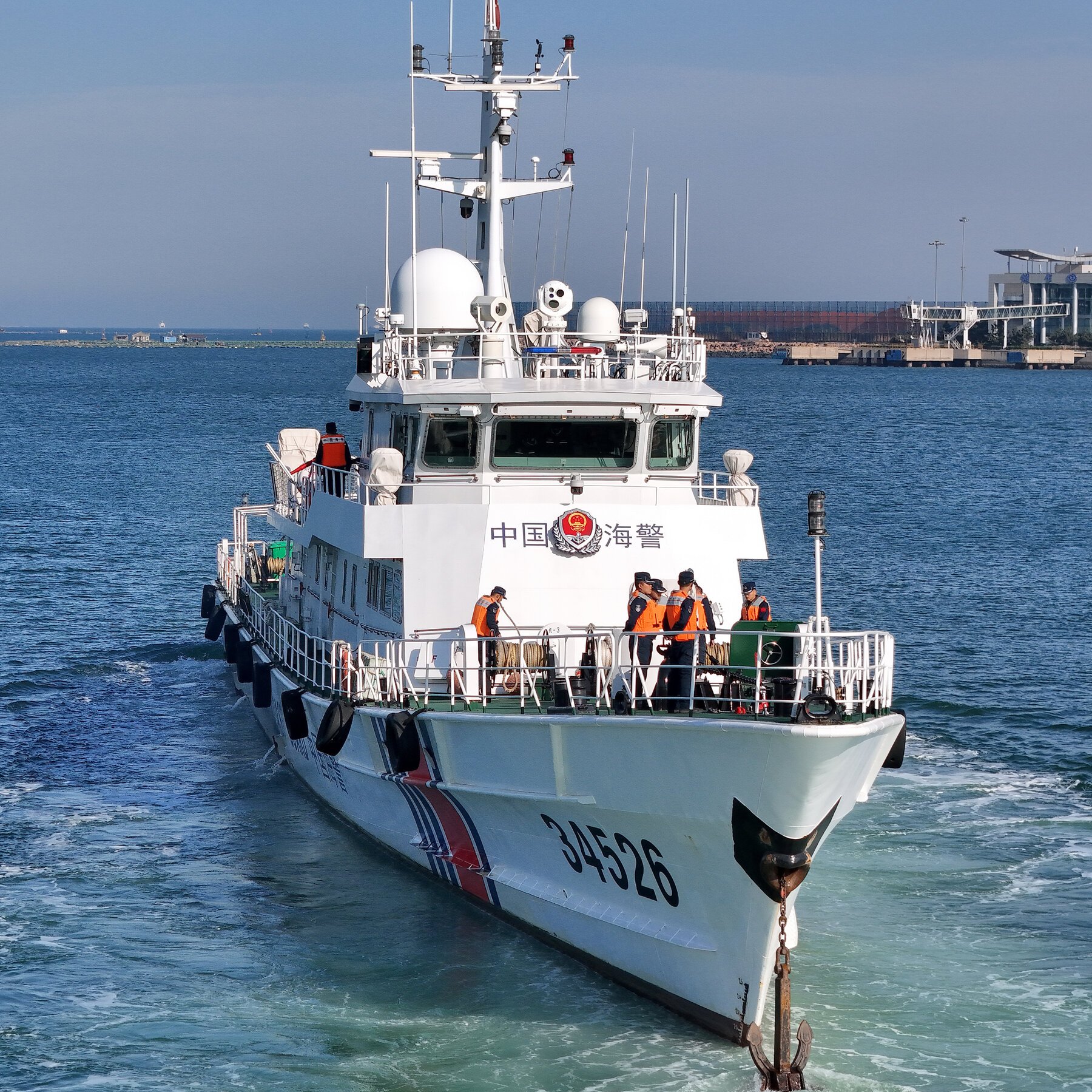 A white patrol vessel marked "34526" with Chinese characters and a crest sails through coastal waters, crew members in orange life vests visible on the bow deck.