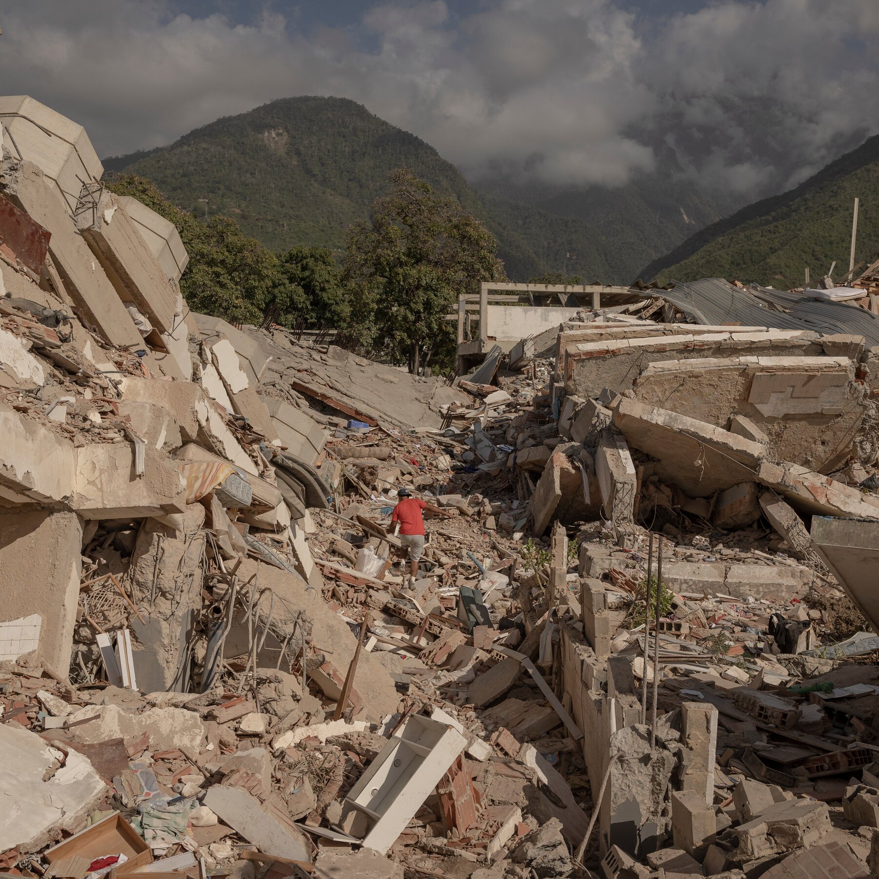 A man in a red shirt walks through extensive concrete and brick building rubble, with forested mountains and low clouds visible in the background.