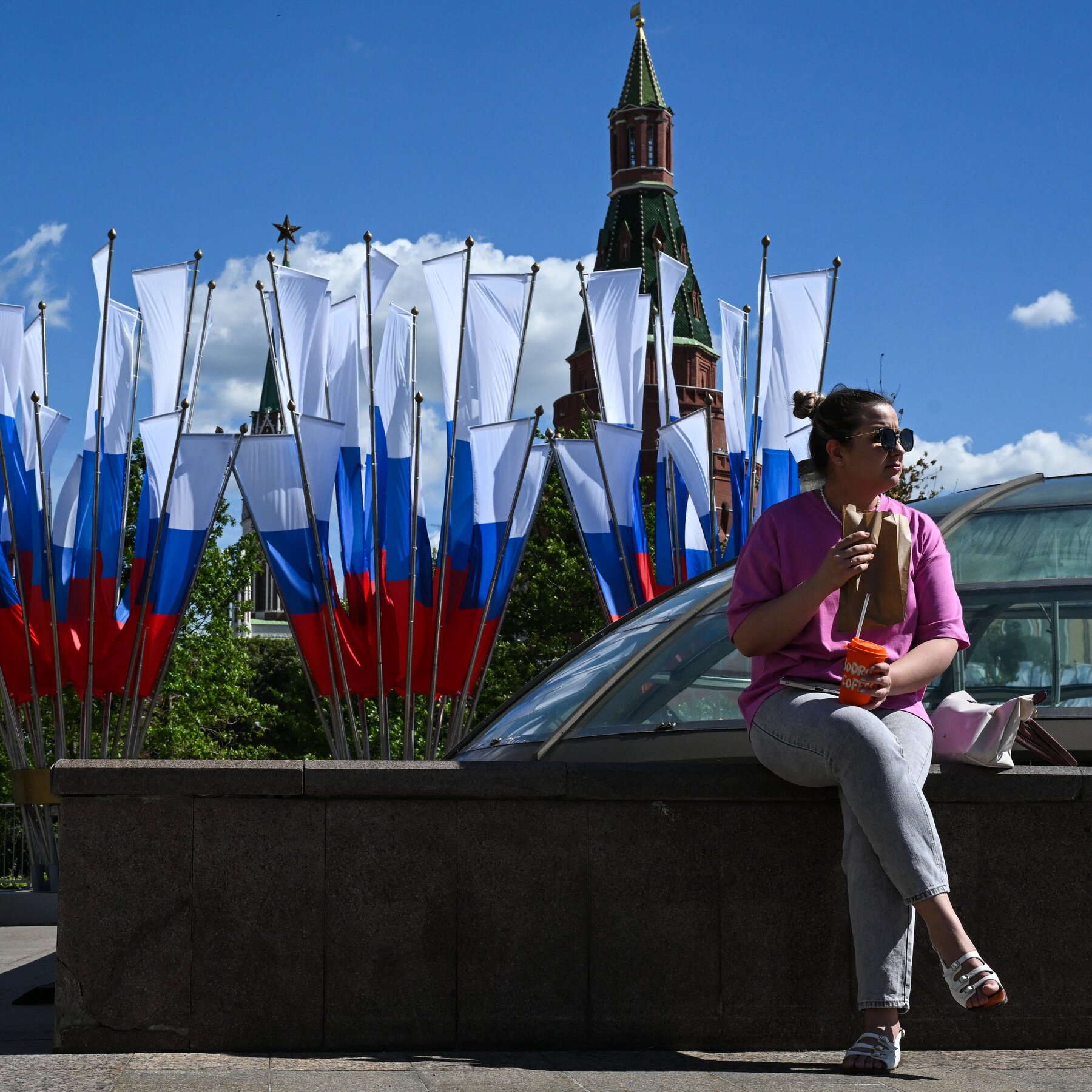 A woman in a pink shirt and sunglasses sits on a stone ledge holding a paper bag and orange drink, with Russian flags and a red-brick tower visible behind her.