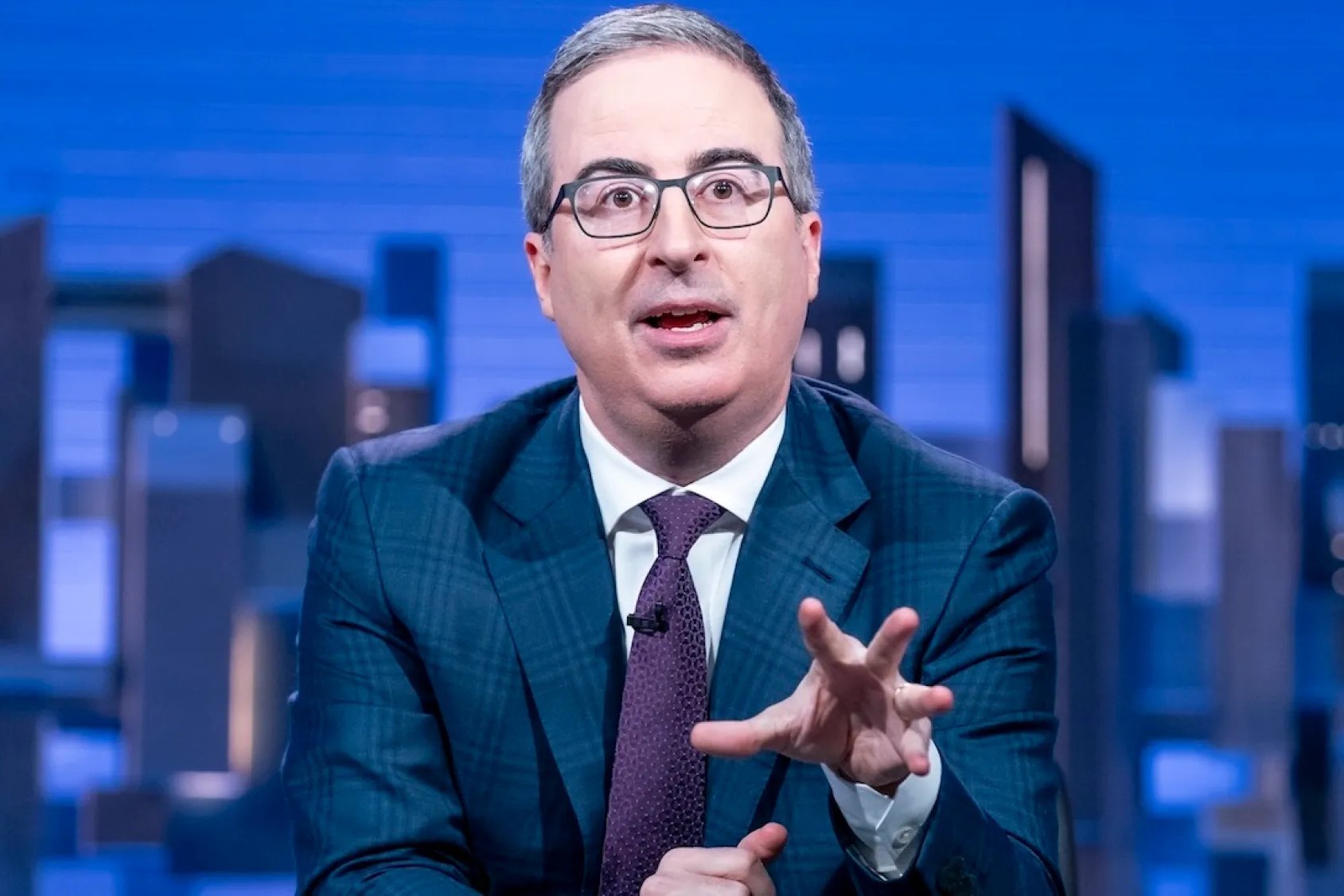 A man in a teal plaid suit and purple tie gestures with his hand while speaking against a blue cityscape backdrop.