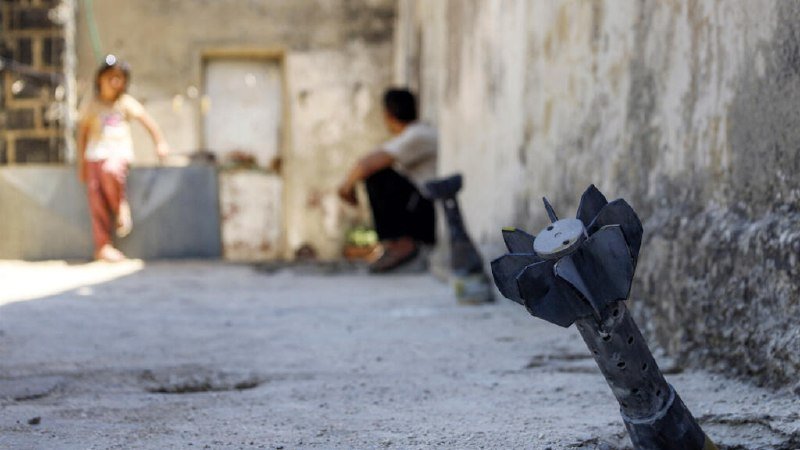 An unexploded ordnance protrudes from the ground in the foreground, while two children sit and stand near weathered buildings in the blurred background.