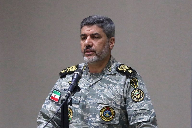 A gray-bearded military officer in camouflage uniform with insignia and an Iranian flag patch speaks into a microphone against a plain beige backdrop.