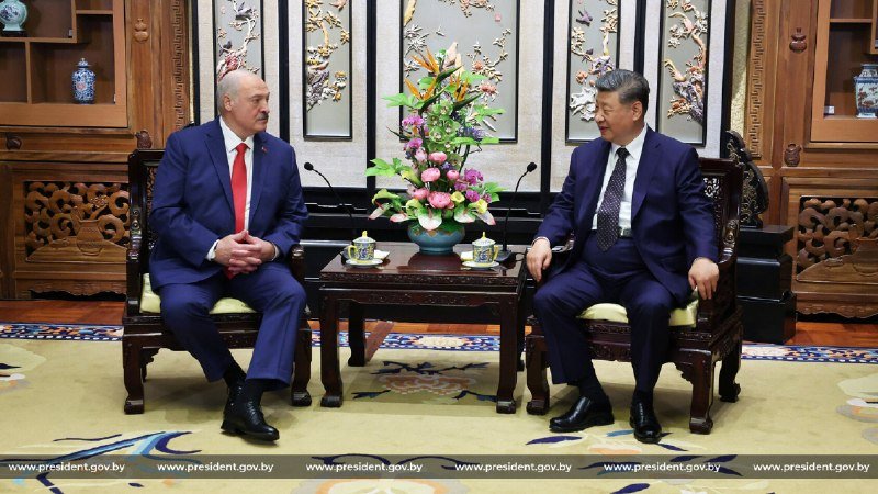 Two men in dark suits sit facing each other in ornate wooden chairs, separated by a small table with a floral arrangement, in a room with decorative screens.
