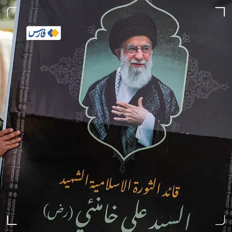 A hand holds a black poster featuring a portrait of a turbaned, white-bearded man with his hand over his chest, above Arabic script.