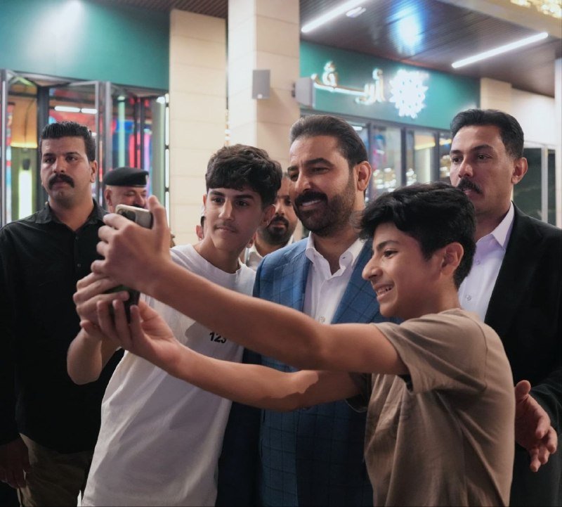 A young boy takes a selfie with a smiling bearded man in a blue blazer, surrounded by several other men outside a storefront with Arabic signage.