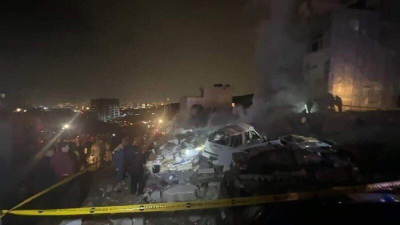 Emergency responders work at night amid debris and a damaged vehicle behind yellow caution tape, with city lights and smoke visible in the background.