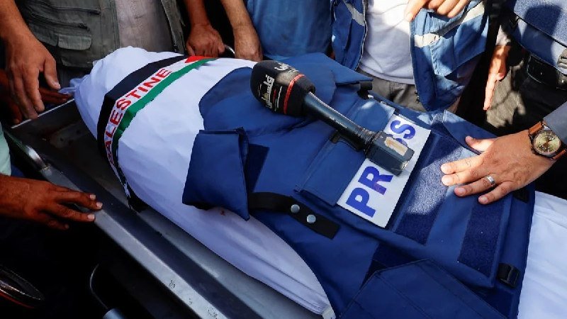 A body wrapped in a Palestinian flag with a "PRESS" vest and a microphone rests on a stretcher, surrounded by several people.