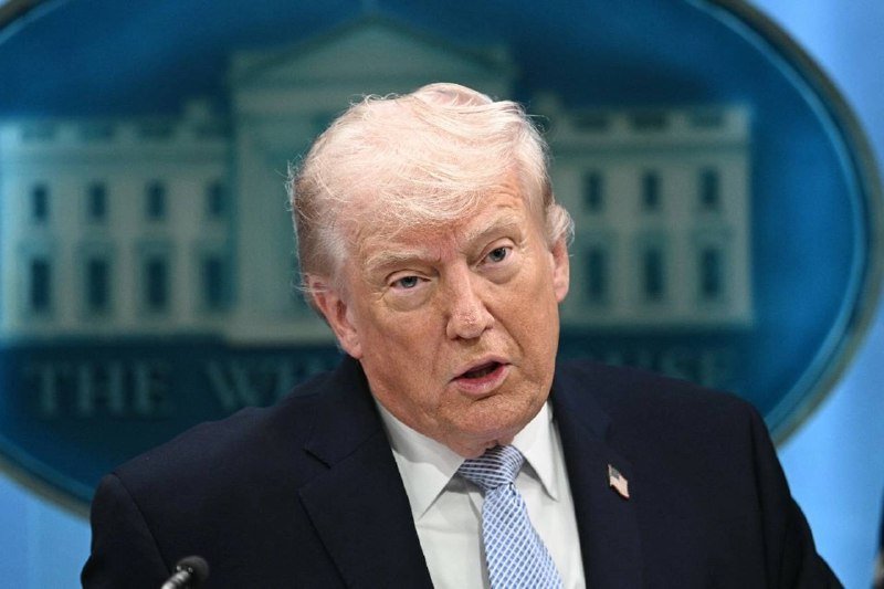 A blonde-haired man in a dark suit and light blue tie speaks at a podium with a microphone, backed by a blue seal bearing a white building illustration and partial text reading "THE WH..."