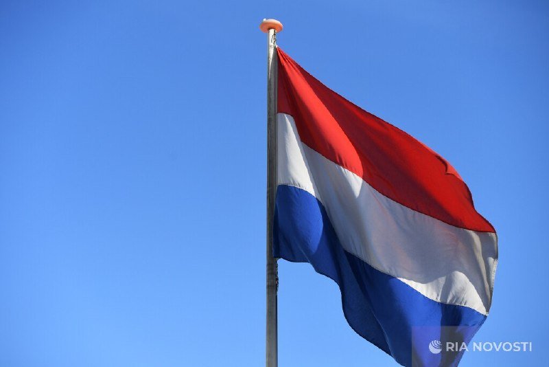 A red, white, and blue horizontal-striped flag waves on a flagpole against a clear blue sky, with a "RIA Novosti" watermark visible in the corner.