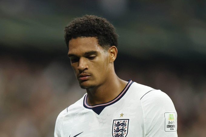 A soccer player wearing a white England national team jersey with a gold star above the crest looks downward during a match, with a blurred crowd visible in the background.