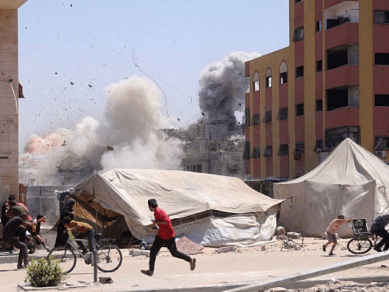 People flee past tents near a damaged multi-story building as smoke billows from a large explosion in a dusty urban street.