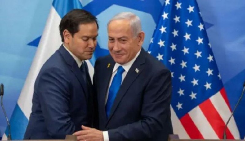 Two men in dark suits stand before Israeli and American flags, smiling and shaking hands at a podium with microphones.