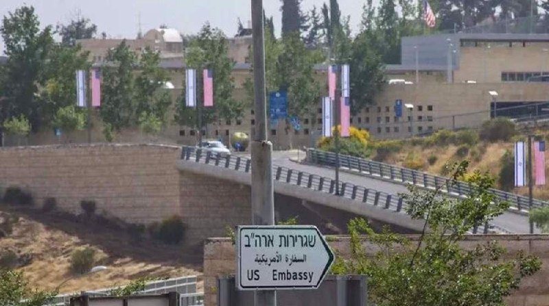 A directional sign in Hebrew, Arabic, and English reads "US Embassy," pointing toward a walled compound with pink banners, security barriers, and a distant American flag.