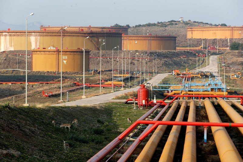 Large orange storage tanks marked "608" stand on a hillside, connected to a network of red and yellow pipelines in the foreground, with antelopes grazing nearby.