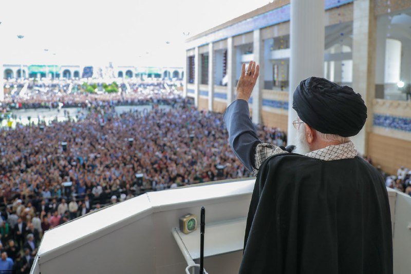 A cleric in black robes and turban waves toward a massive crowd gathered in an open courtyard during what appears to be a religious observance.