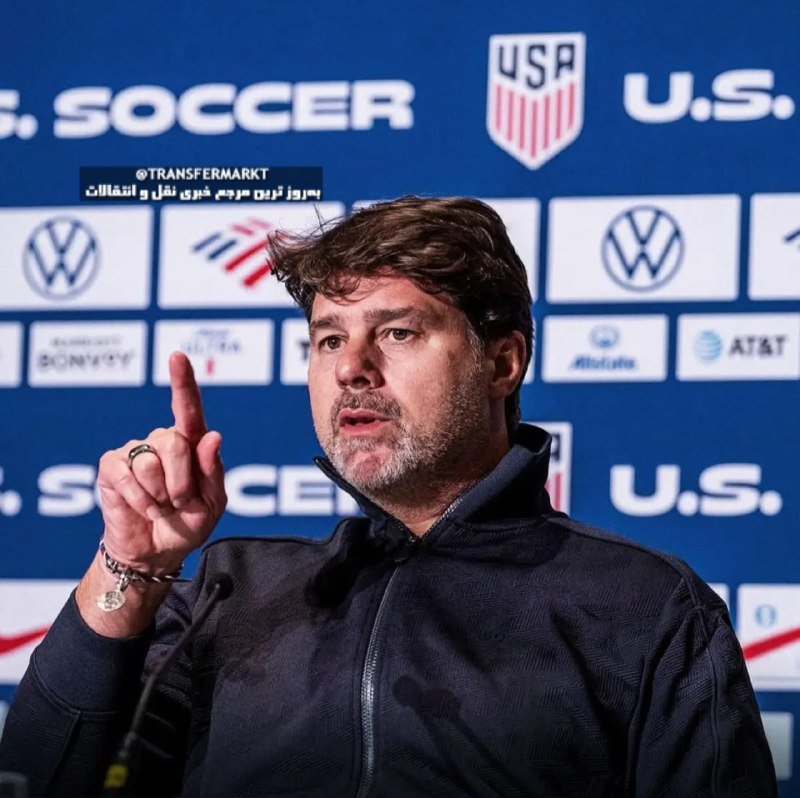 A bearded man in a black jacket gestures with his index finger raised while speaking at a press conference against a backdrop featuring U.S. Soccer and sponsor logos.