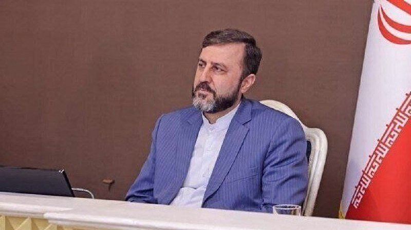 A bearded man in a blue pinstripe suit sits at a table beside an Iranian flag.