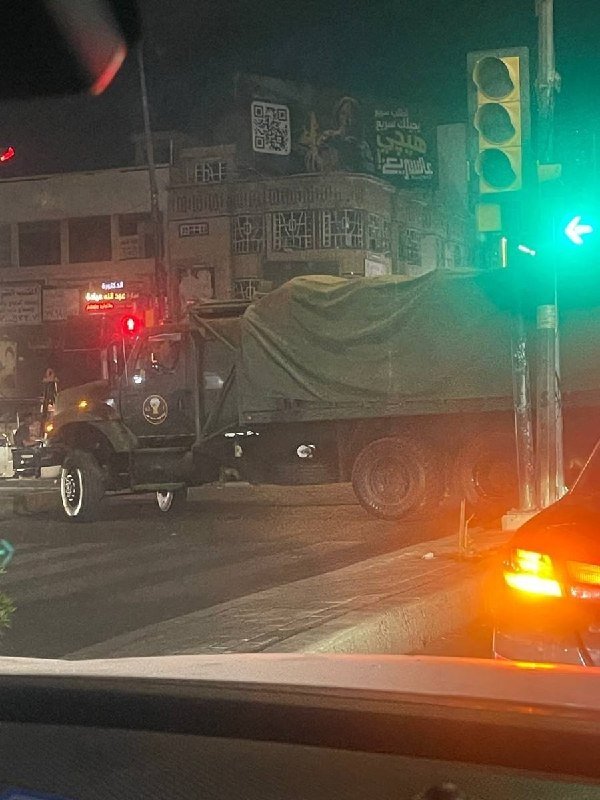 A tarp-covered military-style truck waits at a nighttime intersection under a green traffic light, with Arabic storefront signage visible in the background.