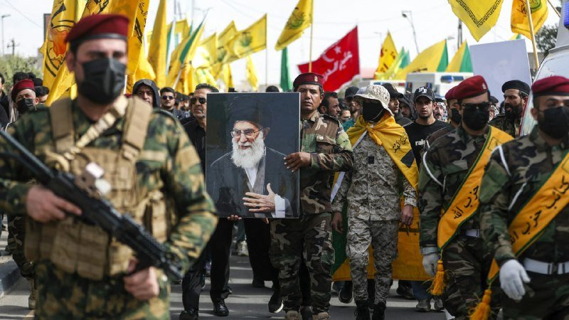 Armed men in military camouflage and red berets march down a street, carrying a large framed portrait of a bearded cleric in black robes while waving yellow flags.