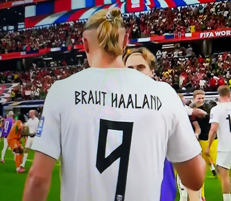 A blonde soccer player wearing a white jersey with "BRAUT HAALAND" and the number 9 stands on a field in a packed stadium.