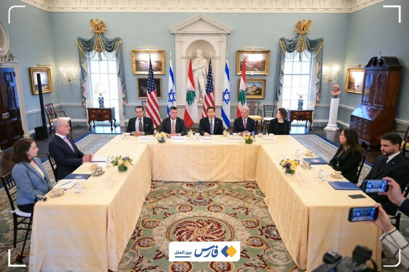 Officials in business attire sit around a U-shaped conference table in an ornate room, with U.S., Israeli, and Lebanese flags displayed behind them.