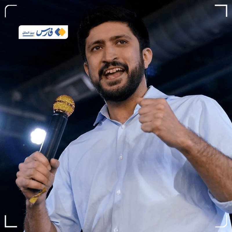 A bearded man in a light blue shirt speaks into a gold-topped microphone, raising his fist while addressing an unseen audience under bright stage lighting.