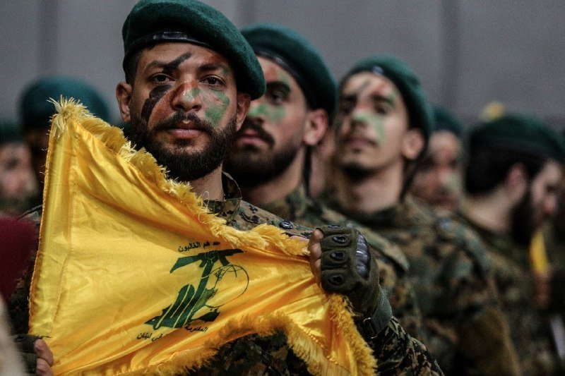 Bearded men in camouflage uniforms and green berets, faces painted in camouflage colors, hold a fringed yellow flag bearing a green emblem and Arabic script.