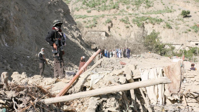 Armed man in camouflage stands among rubble and collapsed structures while a group of civilians gathers in the background of a damaged mountainous village.