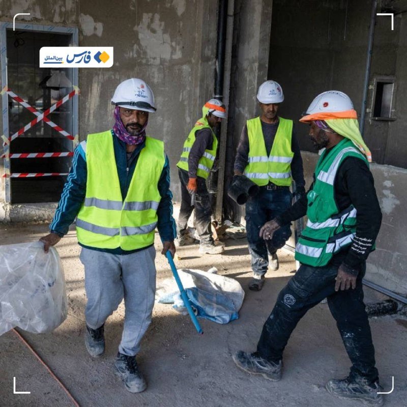 Construction workers in hard hats and high-visibility vests walk through an unfinished concrete building site, carrying bags and tools.