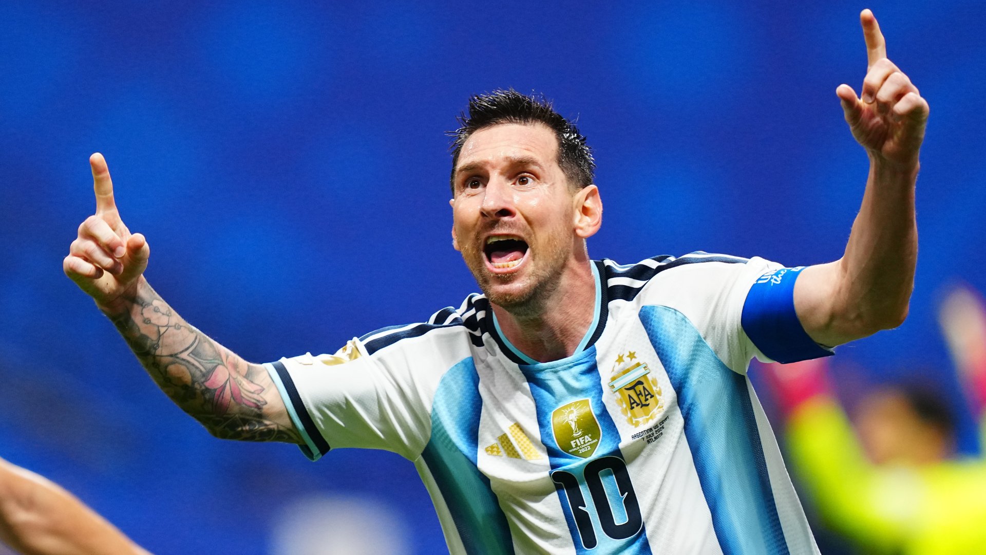 A bearded soccer player wearing a blue and white striped Argentina #10 jersey and a blue captain's armband raises both index fingers toward the sky in celebration against a blue background.