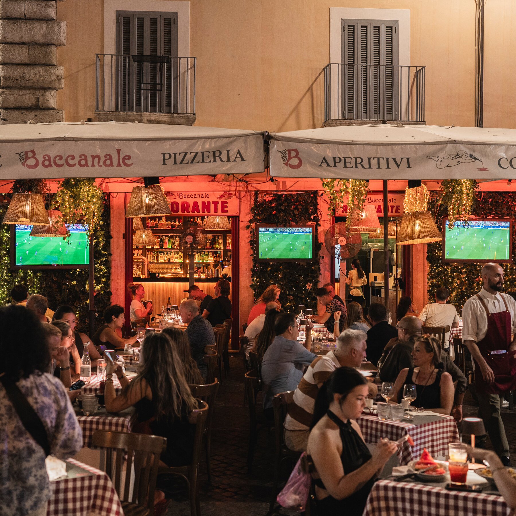 Crowds dine at outdoor tables under white awnings marked "Baccanale Pizzeria" and "Aperitivi," with three televisions broadcasting soccer matches to the seated patrons.