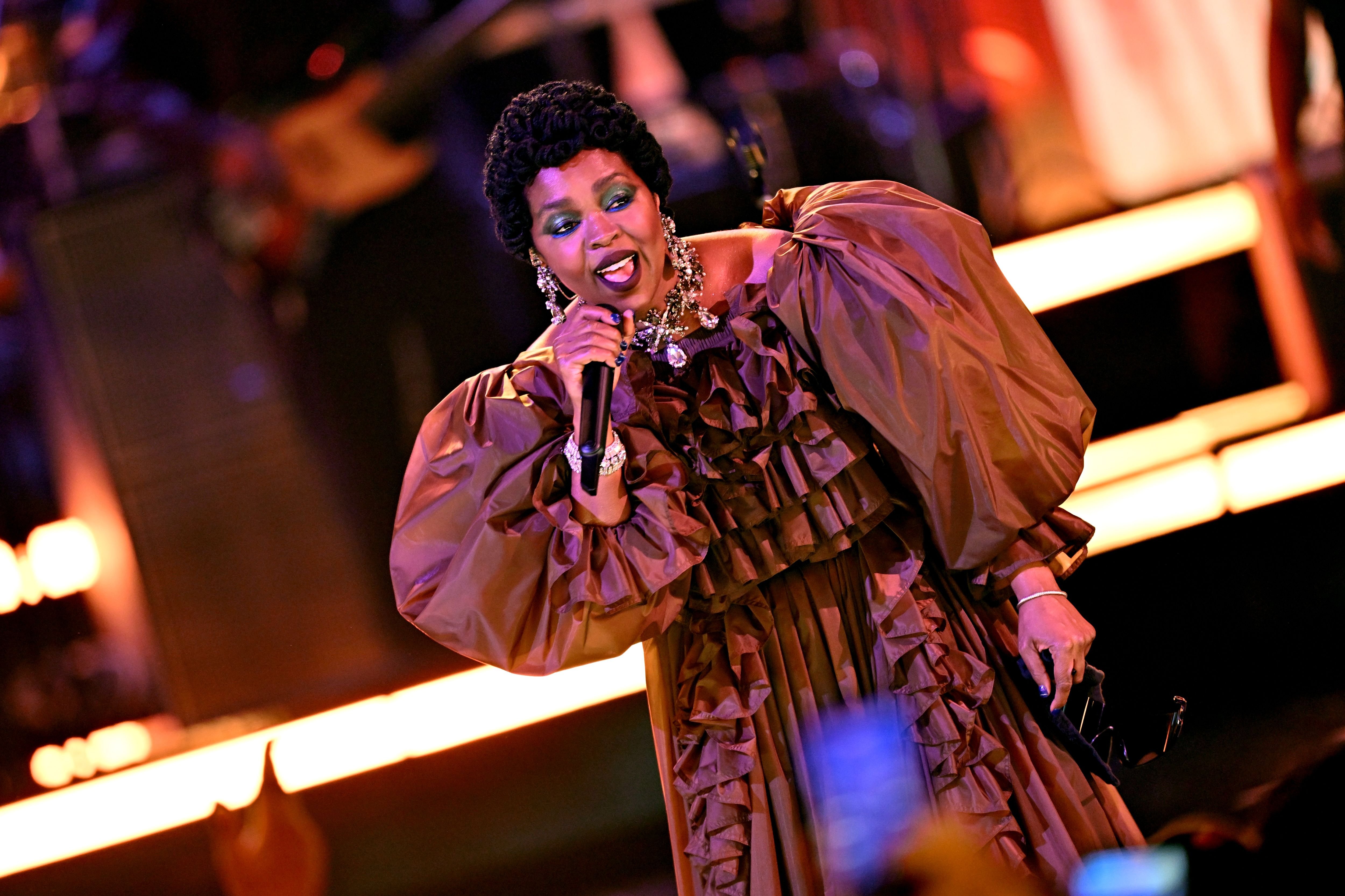 A woman in a ruffled bronze gown with statement jewelry sings into a microphone on a warmly lit stage.
