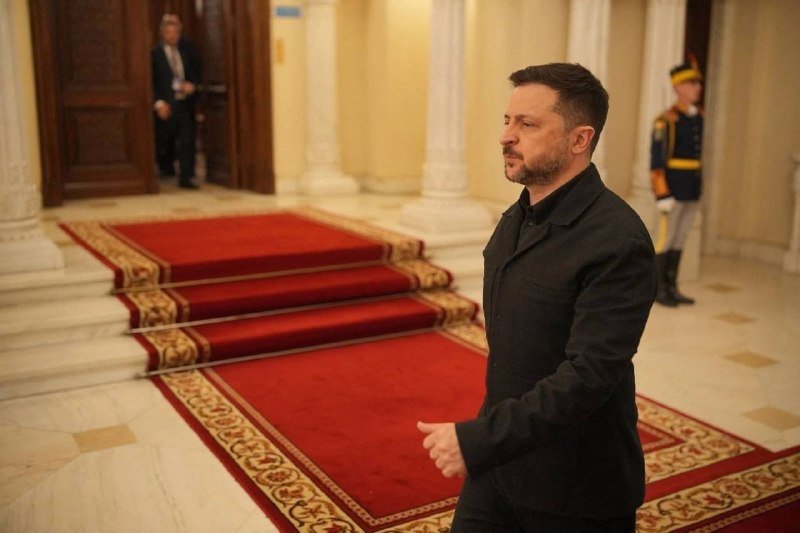 A bearded man in a black coat walks along a red carpet in a grand hall, while a uniformed guard stands at attention in the background.