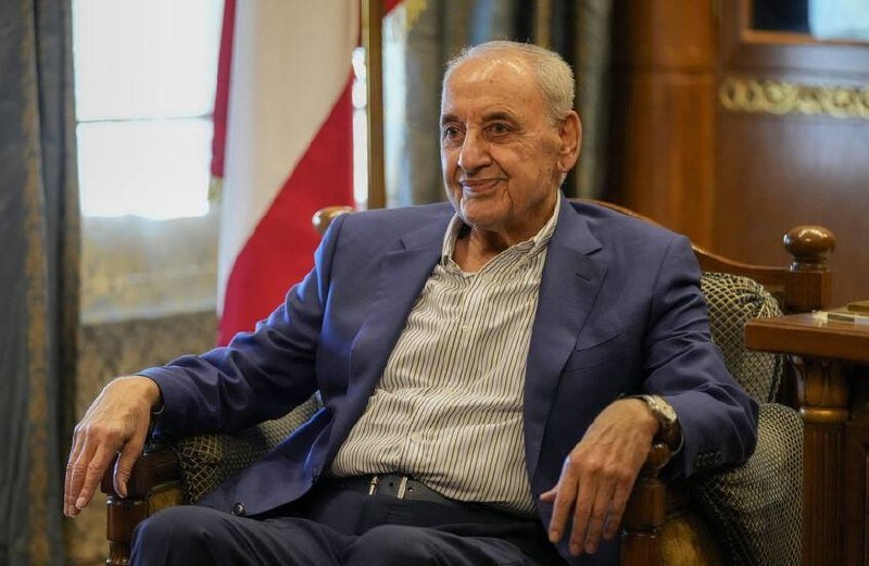 An elderly man in a blue blazer and striped shirt smiles while seated in an ornate chair, with a red-and-white striped flag visible behind him.