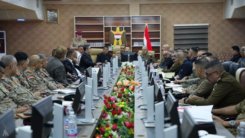 Military officers and officials in uniform sit around a long conference table lined with flowers, monitors, and a flag, during a formal meeting.